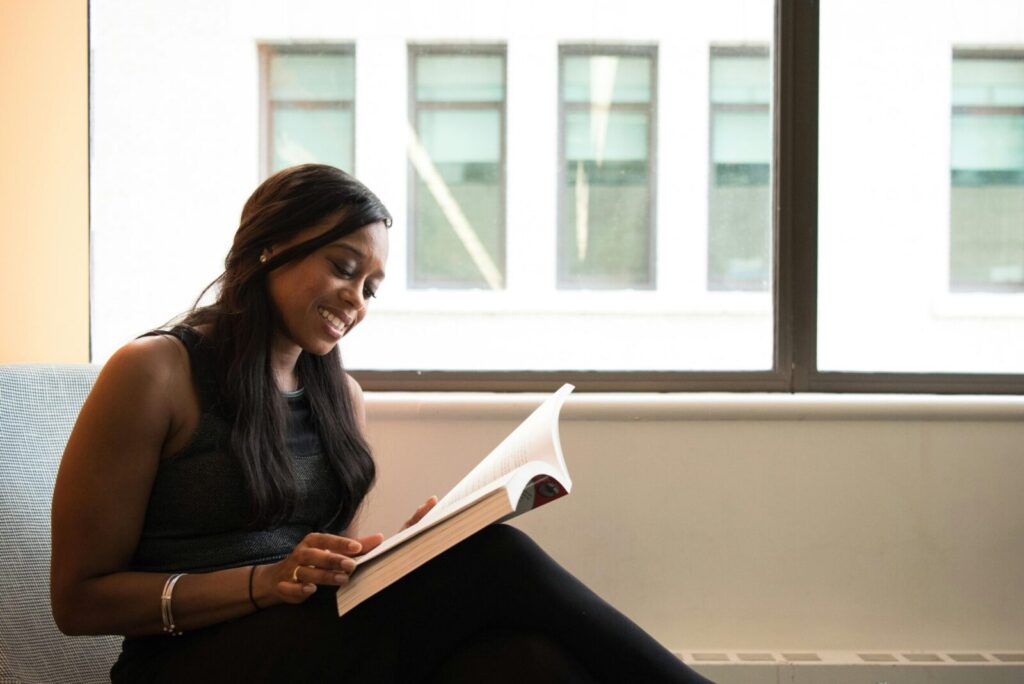 A smiling woman enjoys reading a book indoors by a large window, showcasing leisure and relaxation.