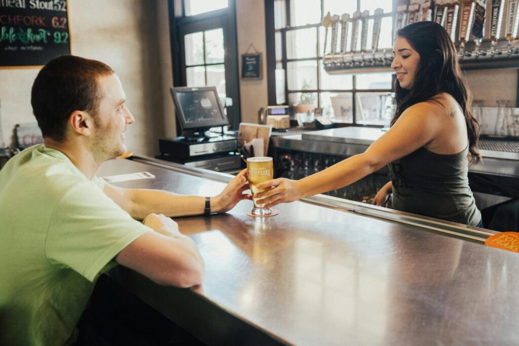 A bartender serves a craft beer to a smiling customer in a lively bar setting.