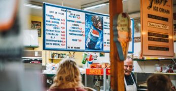 A retro diner setting with a vintage menu board, customers, and a smiling staff member.