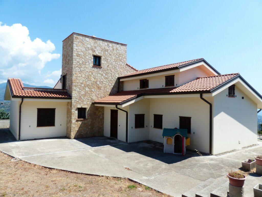 Spacious family house featuring a stone facade and red tile roof, set outdoors under a bright blue sky.