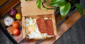 Delicious breakfast box featuring fried egg, sliced meat, and fresh vegetables on a rustic wooden table.