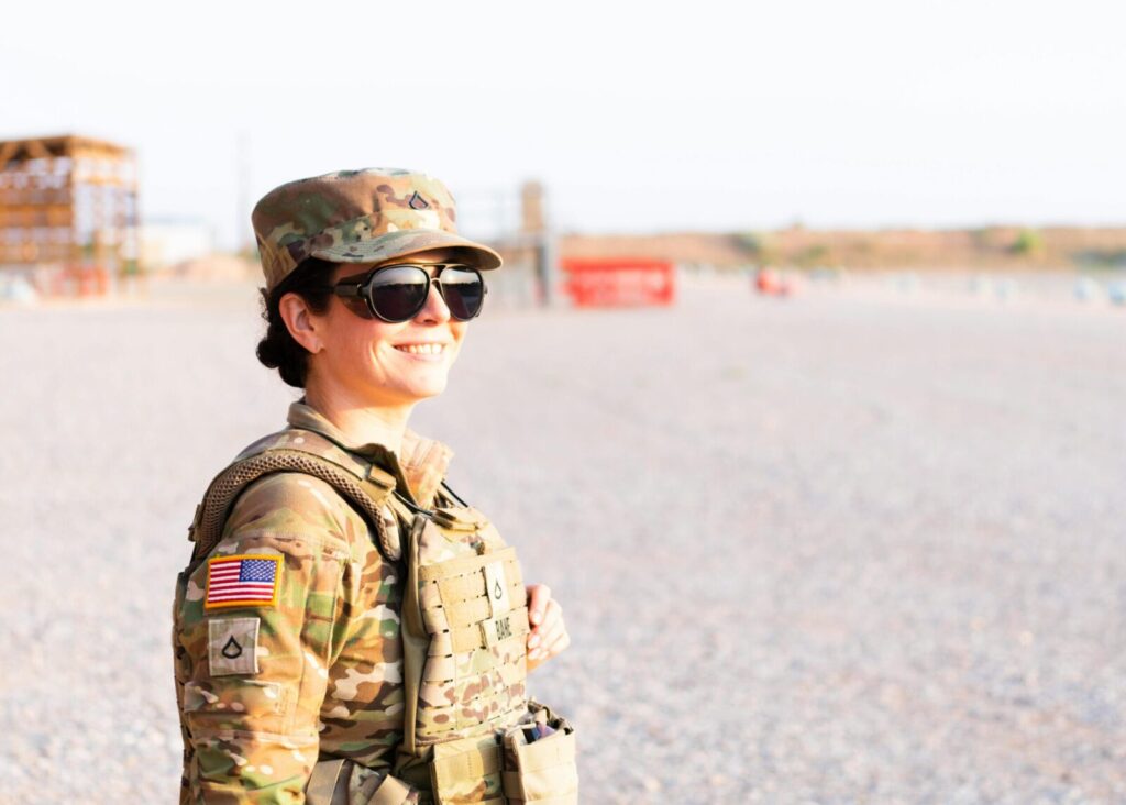 Adult female soldier in camouflage uniform smiling confidently while standing outdoors on a bright day.